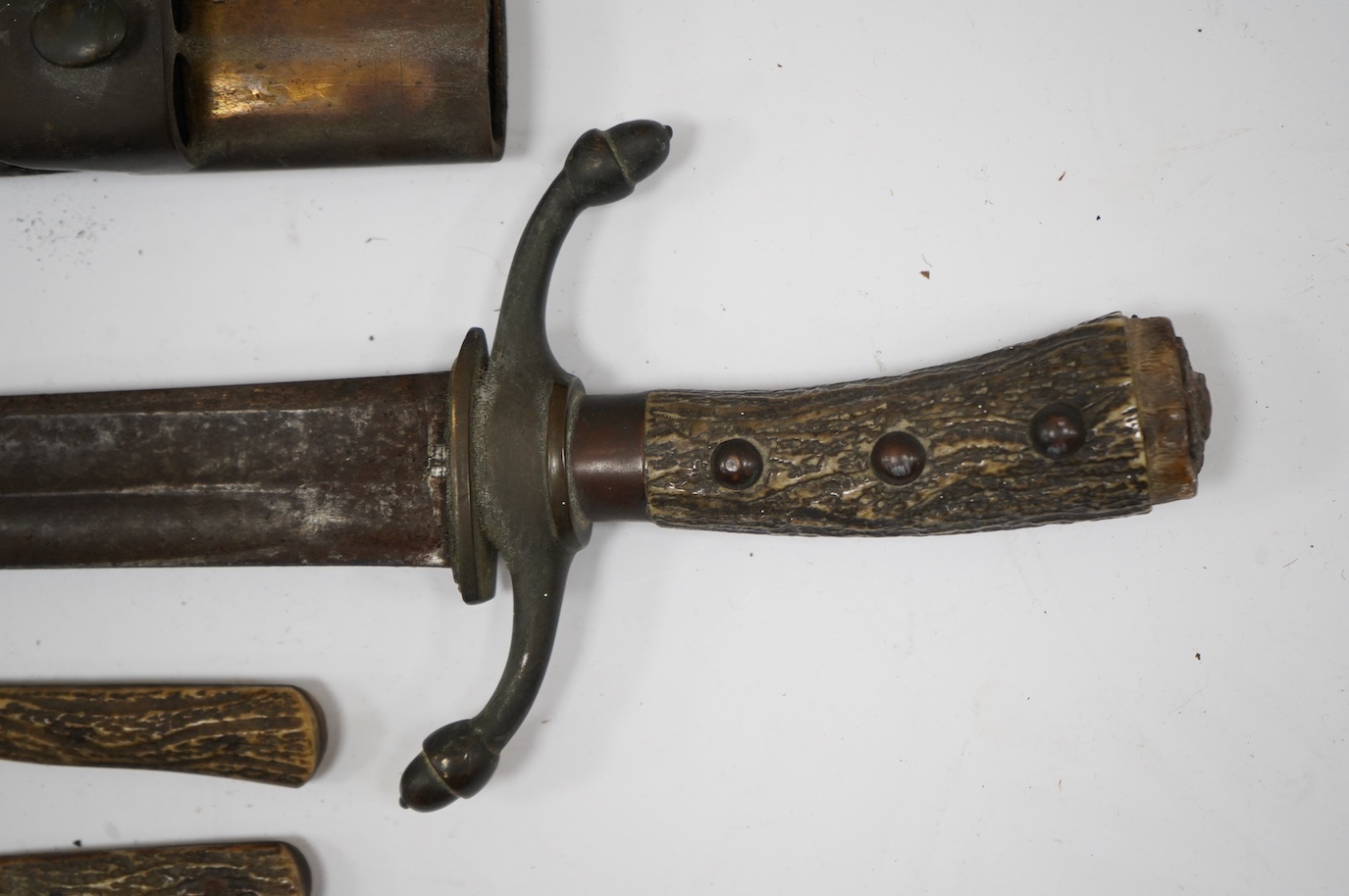 A 19th century Continental hunting hanger, blade cut with twin fullers, recurved brass crosspiece with acorn finials, staghorn grip (pommel missing), in its brass mounted leather scabbard, containing knife and fork (chap